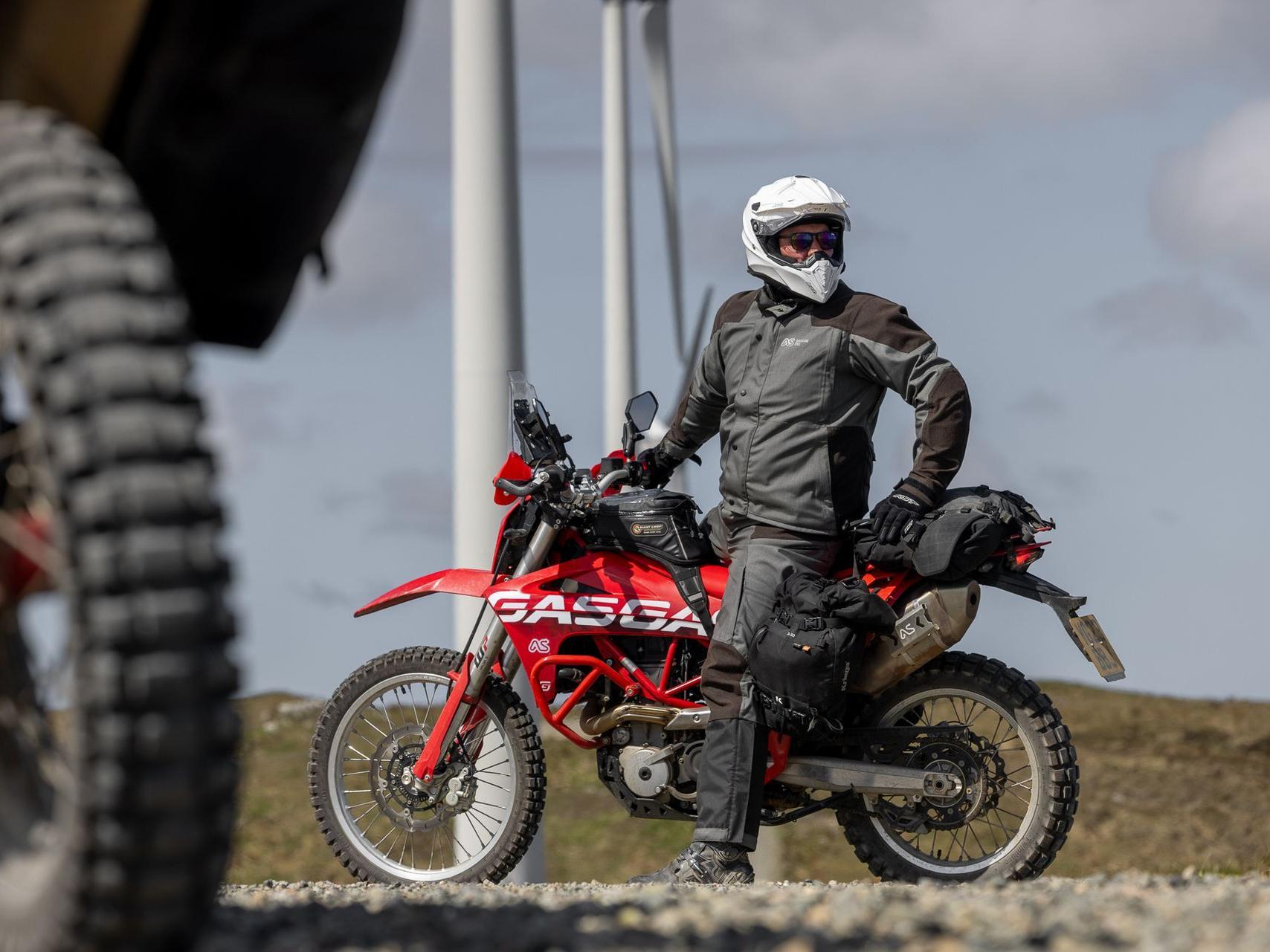 man looking into distance wearing grey gravel jacket and pant sitting on red gasgas es700 nmotorbike with large wind turbines in the background